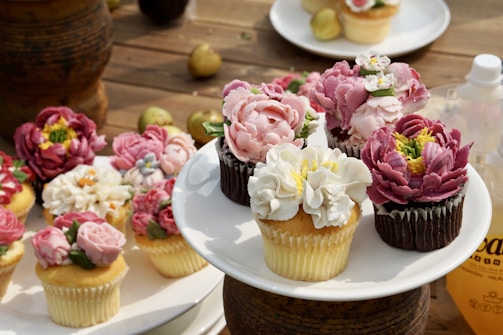 An assortment of cupcakes decorated with intricate, flower-shaped frosting, displayed on a wooden surface. The cupcakes feature a variety of colors, including pink, white, and purple, and are arranged on a white plate. Additional cupcakes are scattered around the main display, and there are some small fruits and a bottle in the background.