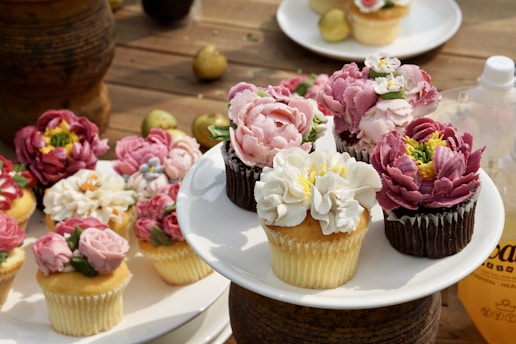 An assortment of cupcakes decorated with intricate, flower-shaped frosting, displayed on a wooden surface. The cupcakes feature a variety of colors, including pink, white, and purple, and are arranged on a white plate. Additional cupcakes are scattered around the main display, and there are some small fruits and a bottle in the background.