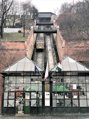 A funicular railway extends up a steep hill, with tracks flanked by brick walls. At the bottom, a glass and iron structure with the sign 'BUDAVARI SIKLO' stands, featuring multiple flags. Trees and sparse vegetation are visible on either side of the tracks.