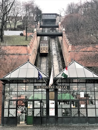 A funicular railway extends up a steep hill, with tracks flanked by brick walls. At the bottom, a glass and iron structure with the sign 'BUDAVARI SIKLO' stands, featuring multiple flags. Trees and sparse vegetation are visible on either side of the tracks.