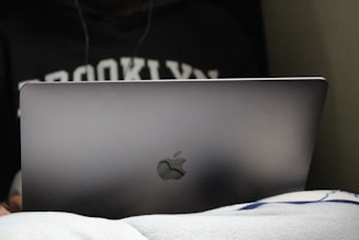 A person is using a laptop, partially covered by a blanket, with the word 'Brooklyn' visible on their sweatshirt.
