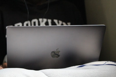 A person is using a laptop, partially covered by a blanket, with the word 'Brooklyn' visible on their sweatshirt.