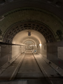 A long, narrow tunnel with an arched ceiling and evenly spaced lights lining the walls. The interior is dimly lit, with greenish-hued tiles on the ceiling and worn concrete walls. The tunnel appears to extend far into the distance, with the floor showing signs of wear and some scattered debris.
