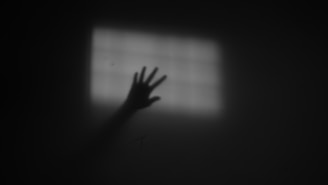 A dark, shadowy hand reaches out towards a softly lit, grid-patterned window, creating an eerie and mysterious atmosphere.