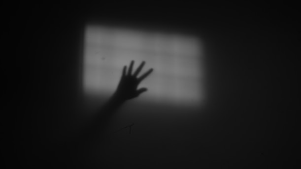 A dark, shadowy hand reaches out towards a softly lit, grid-patterned window, creating an eerie and mysterious atmosphere.