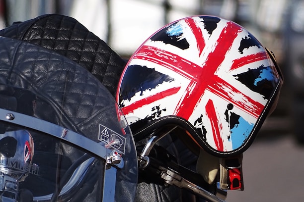 A motorcycle helmet with a Union Jack design rests on a quilted leather motorcycle seat. The helmet features vibrant red, white, and blue colors with a slightly distressed look. Near the helmet is a windshield with reflective surfaces and metal details.