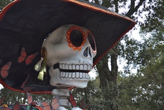 A large skeleton sculpture decorated with traditional colorful patterns, wearing a wide-brimmed hat and adorned with orange butterflies against a backdrop of green trees.