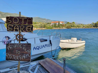 A small wooden sign with rope lettering reads 'Bota Sare' and 'Private Tour to Oyster Farm' next to a dock where a white boat named 'Aquarius' is moored. The background shows a calm body of water with lush greenery and a hill with a building in the distance.