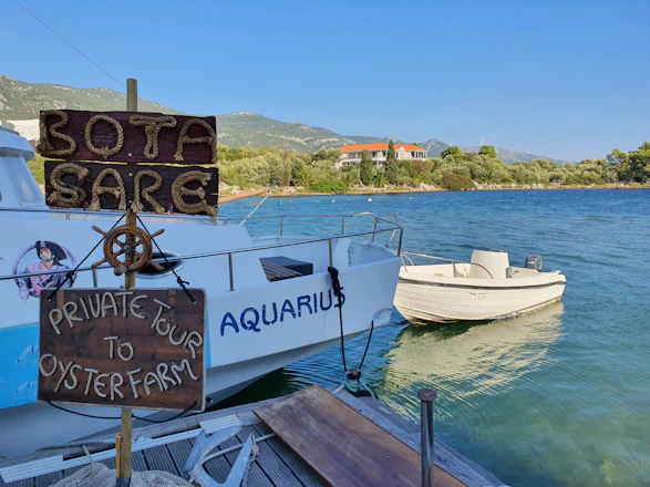A small wooden sign with rope lettering reads 'Bota Sare' and 'Private Tour to Oyster Farm' next to a dock where a white boat named 'Aquarius' is moored. The background shows a calm body of water with lush greenery and a hill with a building in the distance.