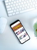 A smartphone displaying a social media or entertainment app, placed on a white desk. Nearby, there is an Apple Magic Keyboard and a white mouse. A partially visible white flower is in the top right corner.