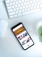 A smartphone displaying a social media or entertainment app, placed on a white desk. Nearby, there is an Apple Magic Keyboard and a white mouse. A partially visible white flower is in the top right corner.