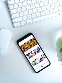 A smartphone displaying a social media or entertainment app, placed on a white desk. Nearby, there is an Apple Magic Keyboard and a white mouse. A partially visible white flower is in the top right corner.