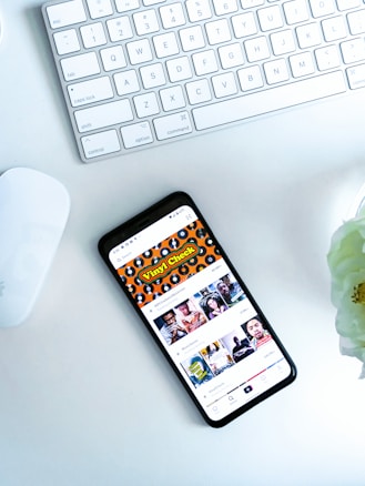 A smartphone displaying a social media or entertainment app, placed on a white desk. Nearby, there is an Apple Magic Keyboard and a white mouse. A partially visible white flower is in the top right corner.