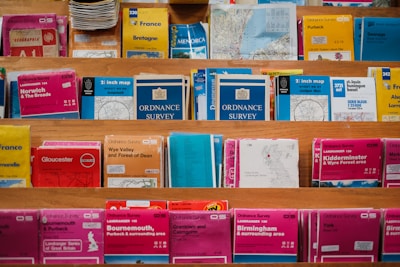 A variety of colorful maps and travel guides are displayed on wooden shelves. The maps are arranged in an orderly manner, with different sizes and titles visible, including Ordnance Survey maps and others for locations like France, Norwich, and Bournemouth.