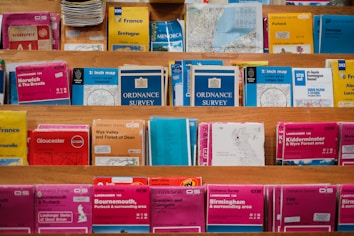A variety of colorful maps and travel guides are displayed on wooden shelves. The maps are arranged in an orderly manner, with different sizes and titles visible, including Ordnance Survey maps and others for locations like France, Norwich, and Bournemouth.