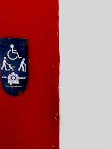 A wall is divided into two sections with a vertical boundary. The left side is a deep red color, while the right side is light gray. Mounted on the red section is a blue sign featuring symbols for accessibility, indicating services for people with disabilities, including figures of a wheelchair, a person with a cane, and a person with a stroller. Below these symbols, the words 'CALL HEAR' and a website link are displayed.