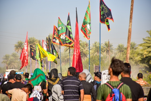 A group of people is gathered outdoors, with many carrying colorful flags, including red, black, yellow, and green with intricate designs. Palm trees are visible in the background, suggesting a warm climate, and the scene conveys a sense of movement or procession.