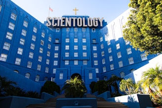 A large, imposing building with a bright blue facade, featuring numerous windows and a prominent sign that reads 'SCIENTOLOGY' in large letters at the top. The building has a cross above the sign and an American flag nearby. It is surrounded by well-maintained greenery, including trees and bushes, creating a welcoming entry path.