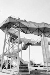 A modern architectural structure featuring a geometric design with multiple platforms connected by staircases. The structure is supported by tall vertical beams, and there are people visible on the upper level. The background is a clear sky, and there are some trees visible at the base.