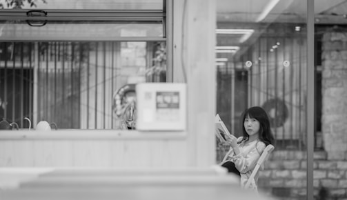 A woman is seated inside a modern, minimalistic space with glass walls, holding and reading a book. The setting seems calm and quiet, with a blend of contemporary and rustic elements, such as glass and stone.