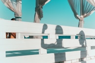 A wooden fence with shadows of two people on it, set against the backdrop of a clear blue sky and ocean. The fence is painted white and has sections that allow light to pass through, creating defined shadows. Curtains tied around columns are visible, adding to a relaxed, beach-side atmosphere.