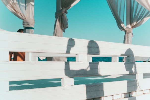 A wooden fence with shadows of two people on it, set against the backdrop of a clear blue sky and ocean. The fence is painted white and has sections that allow light to pass through, creating defined shadows. Curtains tied around columns are visible, adding to a relaxed, beach-side atmosphere.