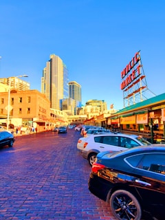 The image depicts an urban street scene with a bustling market area. Tall modern skyscrapers and older brick buildings stand side by side, reflecting a blend of old and new architecture. Cars are parked along a cobblestone street, with people walking nearby. A large red sign reading 'Public Market' is prominently visible against a clear blue sky.