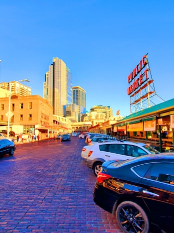 The image depicts an urban street scene with a bustling market area. Tall modern skyscrapers and older brick buildings stand side by side, reflecting a blend of old and new architecture. Cars are parked along a cobblestone street, with people walking nearby. A large red sign reading 'Public Market' is prominently visible against a clear blue sky.