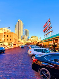 The image depicts an urban street scene with a bustling market area. Tall modern skyscrapers and older brick buildings stand side by side, reflecting a blend of old and new architecture. Cars are parked along a cobblestone street, with people walking nearby. A large red sign reading 'Public Market' is prominently visible against a clear blue sky.