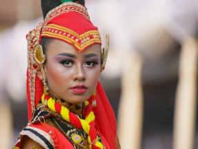 A person is wearing traditional attire with intricate embroidery and vibrant colors. The red headpiece, embellished with gold details, complements the facial makeup featuring bold eyebrows and eyeshadow. The expression is serious and poised, capturing the essence of cultural heritage.
