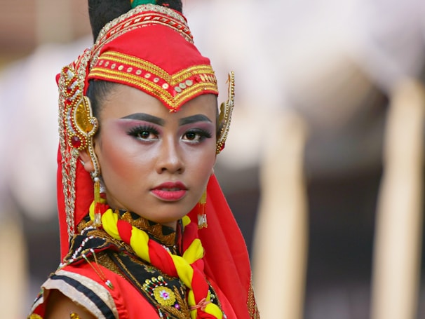 A person is wearing traditional attire with intricate embroidery and vibrant colors. The red headpiece, embellished with gold details, complements the facial makeup featuring bold eyebrows and eyeshadow. The expression is serious and poised, capturing the essence of cultural heritage.