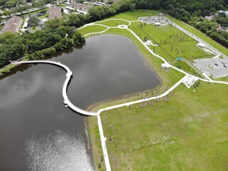 Aerial view of a large, landscaped park featuring a serpentine walkway over a body of water, surrounded by green grass and sparse trees. The park includes parking areas and small buildings, with pathways connecting various sections. Residential buildings and dense vegetation border the park.