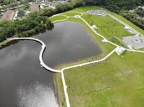 Aerial view of a large, landscaped park featuring a serpentine walkway over a body of water, surrounded by green grass and sparse trees. The park includes parking areas and small buildings, with pathways connecting various sections. Residential buildings and dense vegetation border the park.