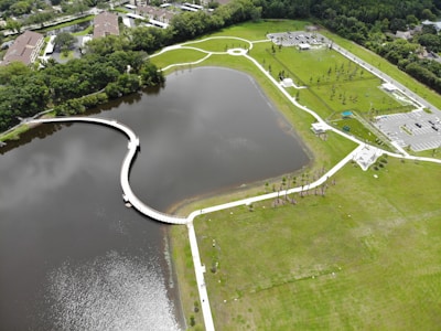 Aerial view of a large, landscaped park featuring a serpentine walkway over a body of water, surrounded by green grass and sparse trees. The park includes parking areas and small buildings, with pathways connecting various sections. Residential buildings and dense vegetation border the park.
