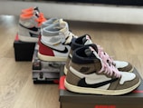 Three pairs of high-top sneakers are placed on shoe boxes, positioned diagonally on a wooden floor. Each pair has distinct color accents, including brown, red, black, and gray colors, with recognizable logos. Laces are untied, and the sneakers appear to be new.