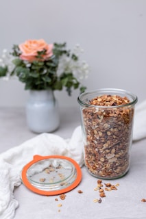 A glass jar filled with granola is placed on a light surface with some granola scattered around. The jar has an orange-rimmed lid lying beside it. In the background, there is a bouquet of flowers in a white vase, featuring a prominent peach-colored rose and green leaves.