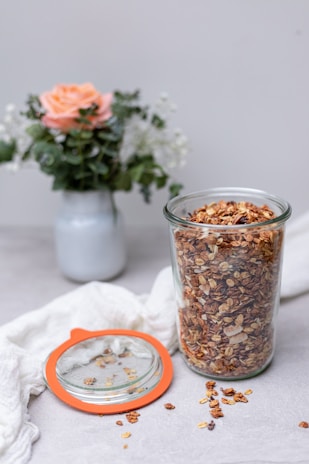 A glass jar filled with granola is placed on a light surface with some granola scattered around. The jar has an orange-rimmed lid lying beside it. In the background, there is a bouquet of flowers in a white vase, featuring a prominent peach-colored rose and green leaves.