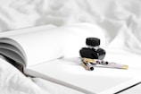 An open notebook with lined pages is placed on a white surface. Beside the notebook is a black ink bottle with a closed lid. Resting on the notebook is an elegant fountain pen with a floral design and a gold-colored cap.