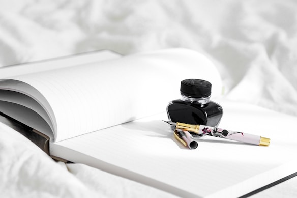 An open notebook with lined pages is placed on a white surface. Beside the notebook is a black ink bottle with a closed lid. Resting on the notebook is an elegant fountain pen with a floral design and a gold-colored cap.