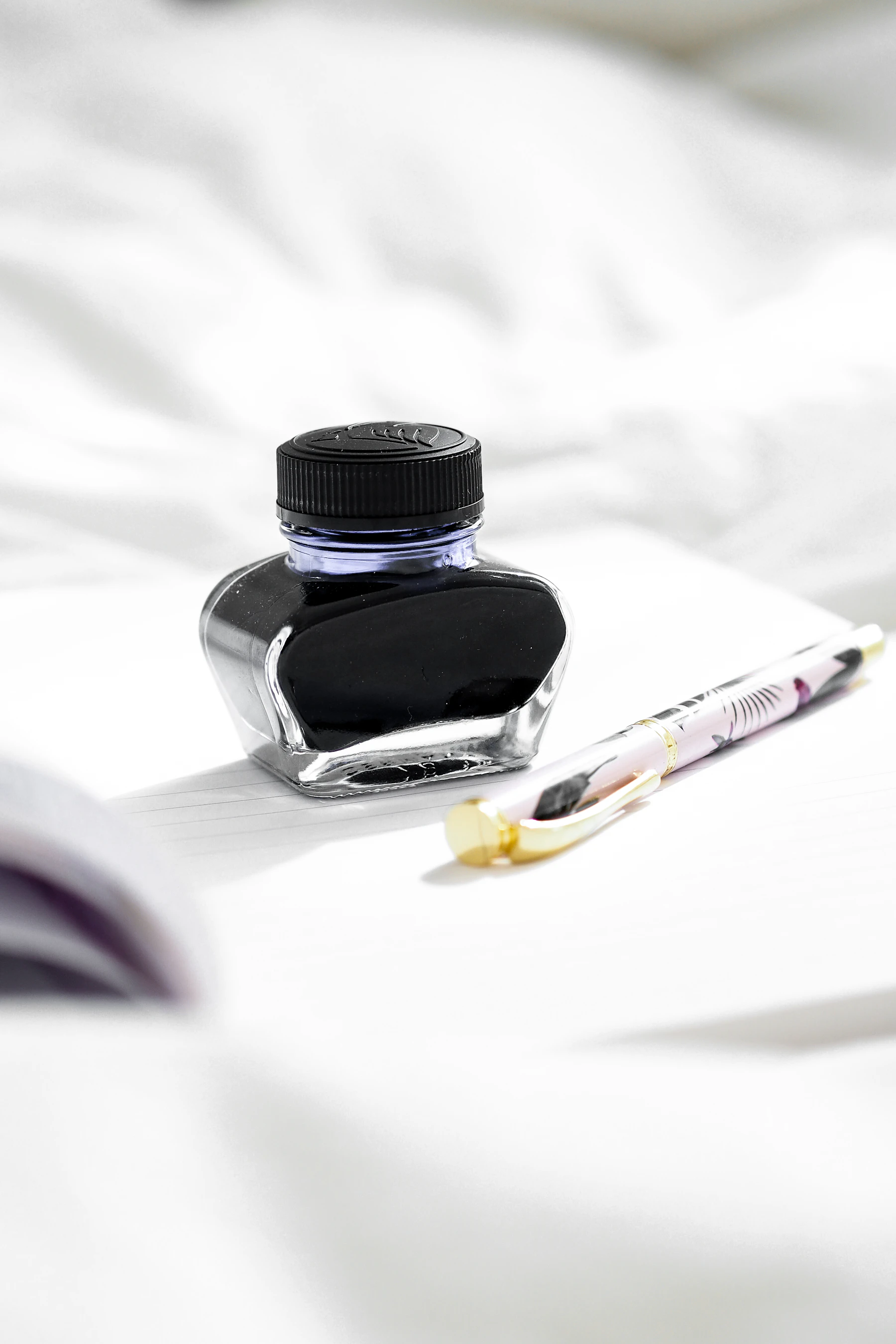 A glass bottle of dark ink beside a clear fountain pen on white paper