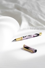 A stylish fountain pen with a floral design and gold accents lies on an open notebook with lined pages. The pen cap is placed nearby on the same surface, and soft natural light illuminates the scene, creating gentle shadows.