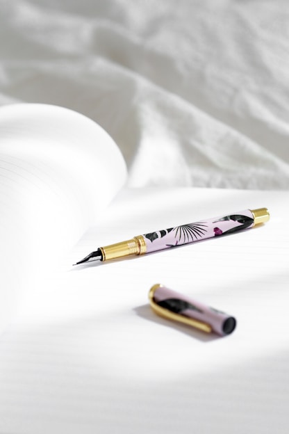 A stylish fountain pen with a floral design and gold accents lies on an open notebook with lined pages. The pen cap is placed nearby on the same surface, and soft natural light illuminates the scene, creating gentle shadows.