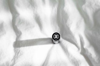 A cylindrical object, possibly a cosmetic product like lipstick, with a branded logo on top, placed on a crumpled white fabric background casting a shadow.