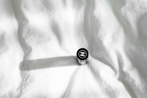 A cylindrical object, possibly a cosmetic product like lipstick, with a branded logo on top, placed on a crumpled white fabric background casting a shadow.