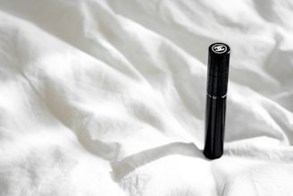 A sleek black cylindrical object with a logo on top is positioned upright on a crumpled white fabric surface. Natural light casts a soft shadow, adding subtle depth to the texture of the fabric.