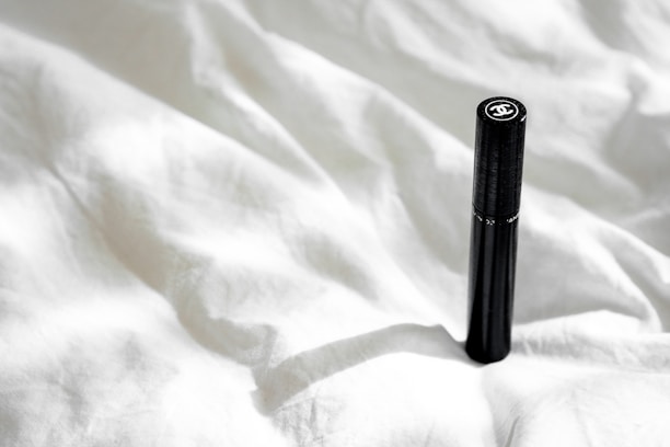 A sleek black cylindrical object with a logo on top is positioned upright on a crumpled white fabric surface. Natural light casts a soft shadow, adding subtle depth to the texture of the fabric.