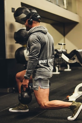 A person wearing a cap and headphones, dressed in workout attire, is performing an exercise using a kettlebell. They are in a gym setting, surrounded by equipment like weight bags. The person is lunging with one leg forward and holding the kettlebell in one hand.