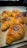 Golden brown croissants with a glossy finish and dollops of chocolate on top are arranged on a wooden board. The pastries have a flaky texture and are displayed in a well-lit setting, suggesting a bakery or kitchen environment.