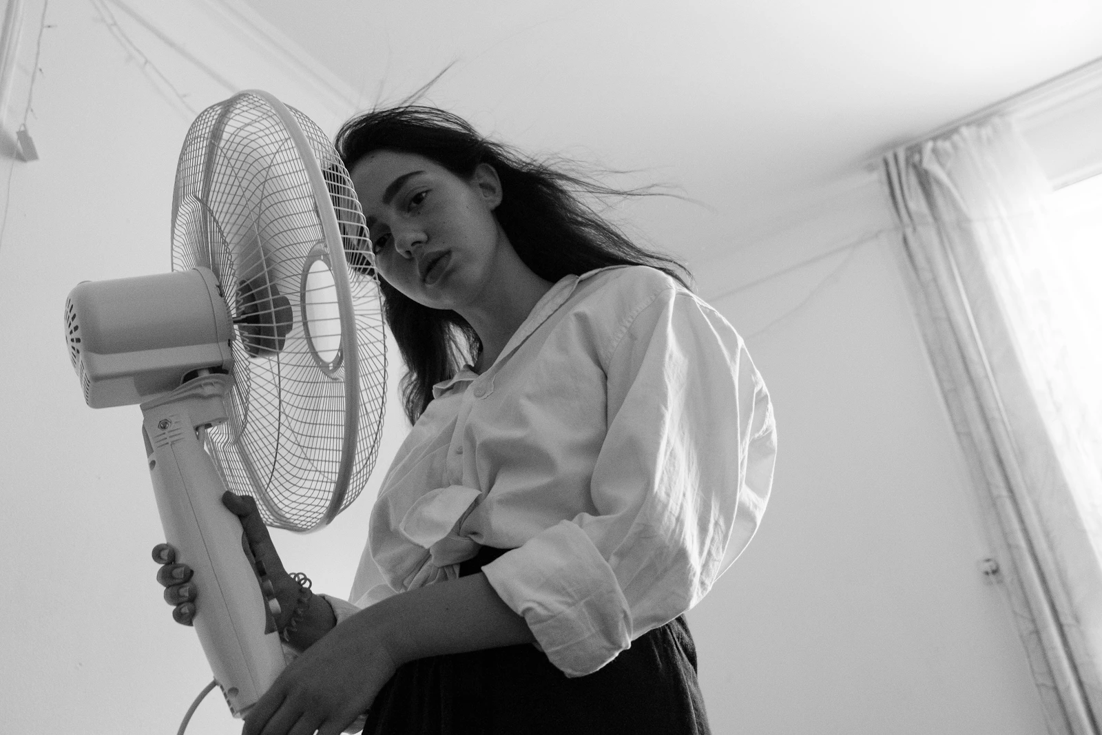 A woman in a white shirt standing beside a large white pedestal fan to stay cool.