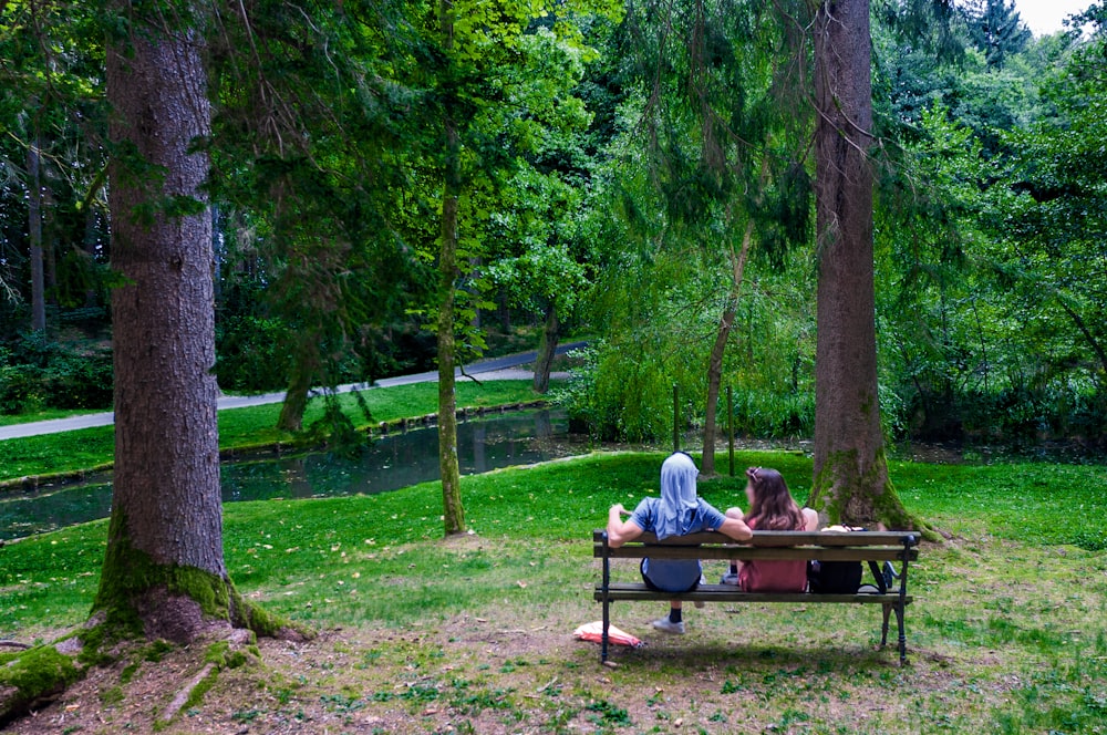 man and woman sitting on brown wooden bench surrounded by green trees during daytime